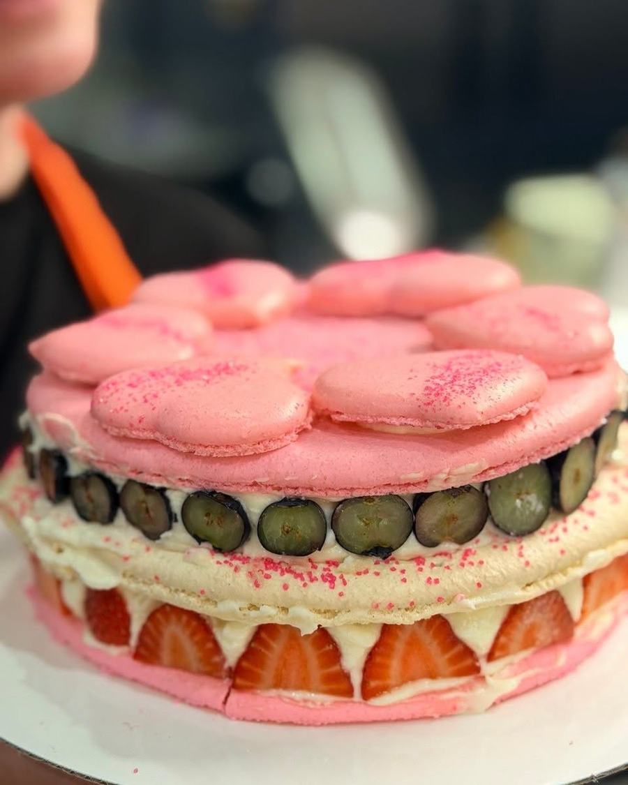 A layered dessert cake topped with pink macarons, fresh grapes and strawberries