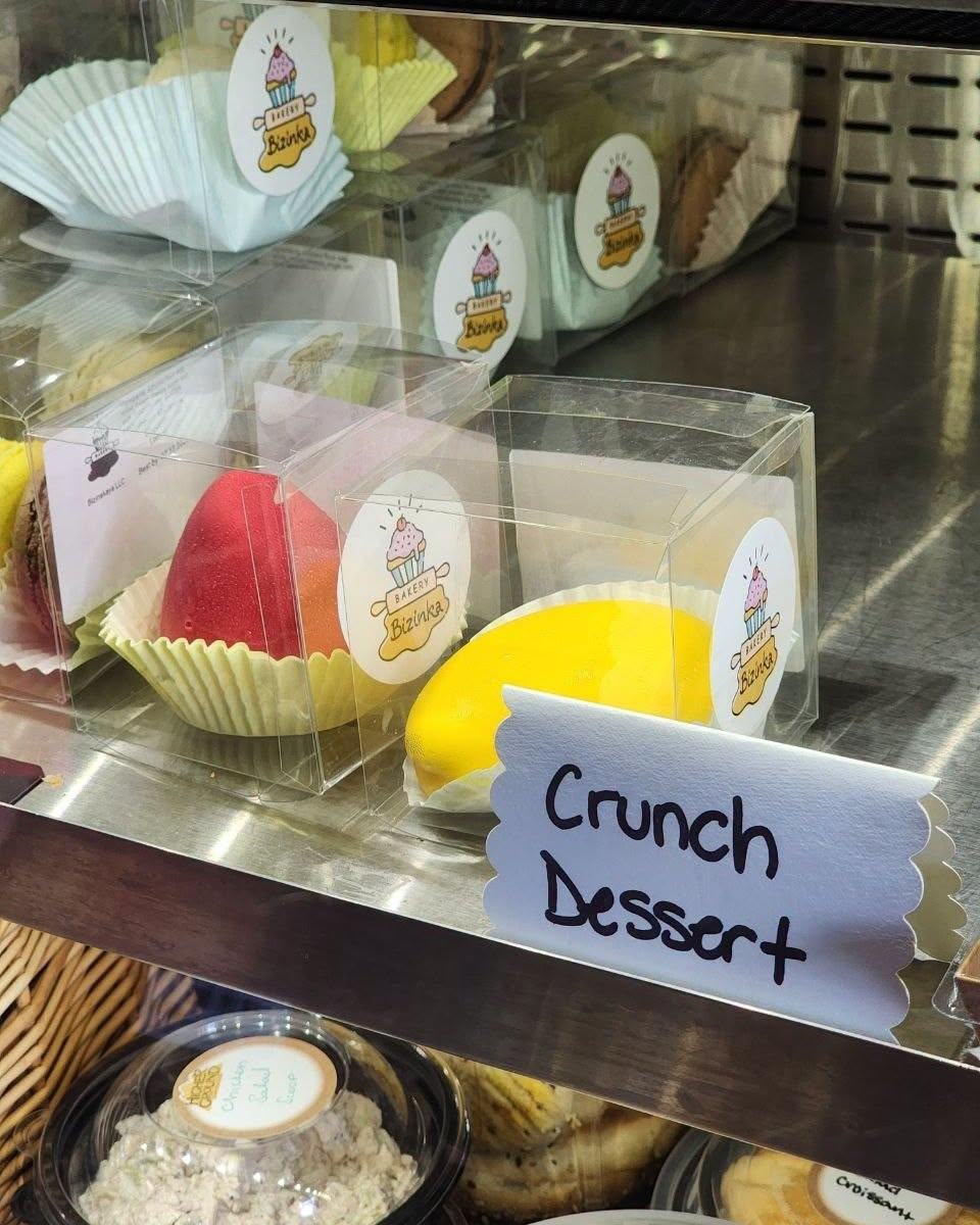 Bizinka Bakery's display case, with mango and berry mousse desserts in branded boxes next to a handwritten "Crunch Dessert" sign
